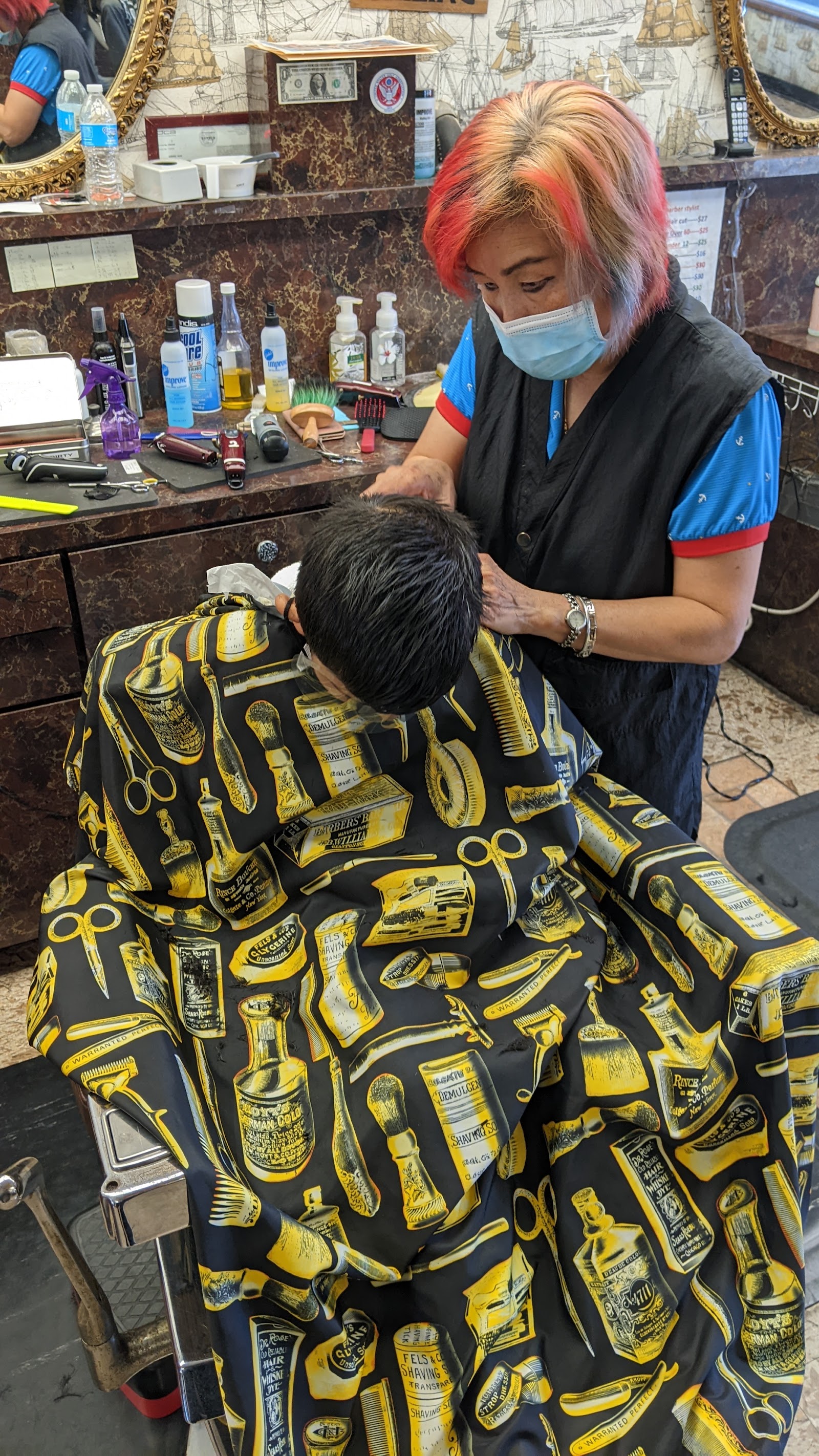 Tina, the lead stylist, cutting a boy's hair. He is wearing a barber cape printed with yellow whiskey bottles and barber tools on black.
